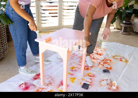 Donna birazziale matura che dipinge sgabello a casa in un progetto upcycling, giovane donna birazziale che assiste Foto Stock