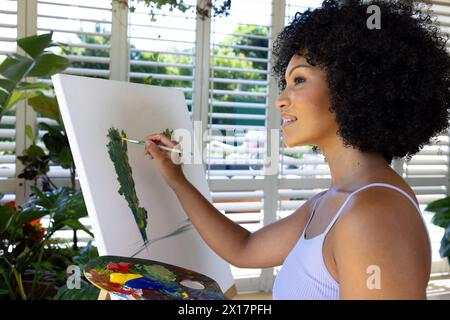 Giovane donna birazziale che dipinge su tela a casa, in piedi accanto alla finestra Foto Stock