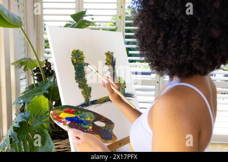 Giovane donna birazziale che dipinge su tela a casa, concentrandosi intensamente Foto Stock
