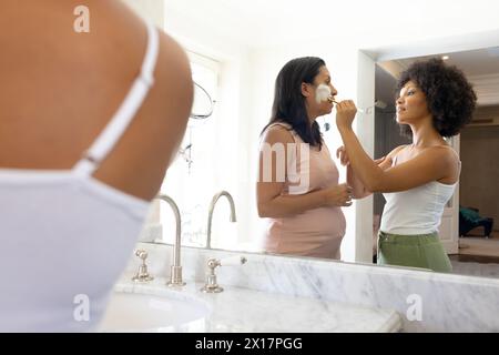 Donna birazziale matura applicazione maschera viso a casa in bagno, giovane donna birazziale guardare Foto Stock