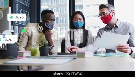 Immagine di icone dei social media su diversi colleghi che indossano maschere facciali che discutono in ufficio Foto Stock