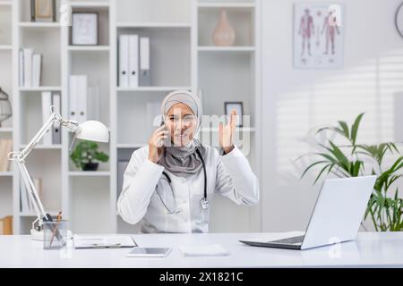 Una dottoressa in un hijab appare stressata mentre parla al telefono e gestiva con l'altra mano in un ufficio moderno ben attrezzato. Foto Stock