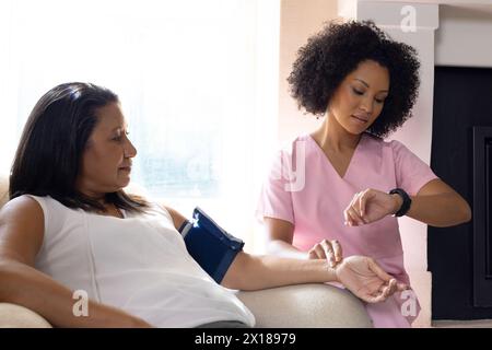 Donna birazziale matura che controlla la pressione sanguigna a casa, giovane infermiera che assiste Foto Stock