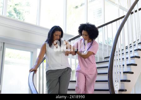 Un'infermiera che assiste una donna asiatica matura giù le scale a casa Foto Stock