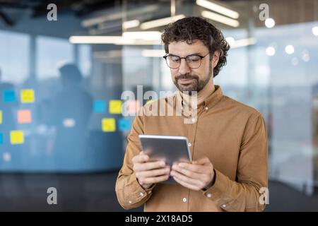 Un professionista specializzato si trova in un ufficio moderno, utilizzando un tablet. Il suo abbigliamento casual e lo sfondo in vetro dell'ufficio con note appiccicose aggiungono un tocco di atmosfera aziendale. Foto Stock