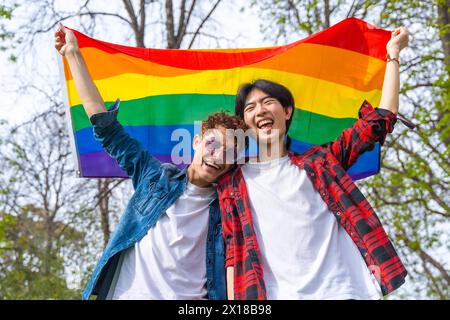 Vista dall'angolo basso ritratto di una coppia gay felice che ride mentre alza la bandiera arcobaleno lgbt all'aperto in un parco Foto Stock