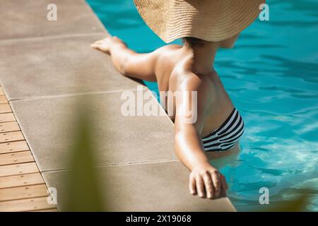 Giovane donna birazziale con cappello appoggiato sul bordo della piscina Foto Stock