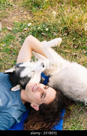 le risate fotografiche verticali riempiono l'aria come un uomo con capelli ricci e il suo simpatico husky condivide un momento di pura beatitudine, disteso sull'erba togeth Foto Stock