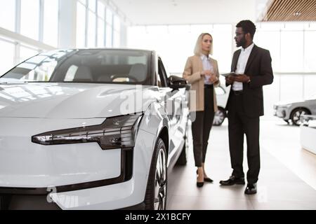 Il consulente maschile africano fornisce informazioni sulle nuove auto tramite tablet a una cliente caucasica presso la concessionaria. Donna d'affari di successo in auto Foto Stock