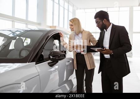 Donna d'affari di successo nello showroom di automobili per la vendita di veicoli ai clienti. Il consulente maschile africano fornisce informazioni sulla nuova auto utilizzando tablet a Cau Foto Stock