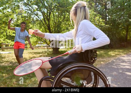 Una donna che usa una sedia a rotelle per partecipare a una partita di badminton con un amico maschio in un parco lussureggiante e soleggiato. Cattura la gioia e lo stile di vita attivo. Foto Stock