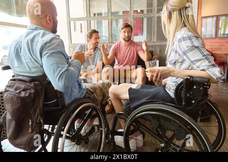 Un gruppo di amici, tra cui persone su sedia a rotelle, si è impegnato in una conversazione vivace e si è gustato un drink insieme in un ambiente soleggiato da caffè. Foto Stock