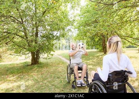 Un ambiente gioioso in un parco dove un uomo e una donna, entrambi in sedia a rotelle, condividono un momento di spensieratezza con un pallone da calcio, raffigurando l'inclusione e un Foto Stock