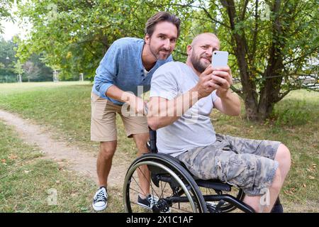 Due uomini che condividono un momento, con uno che usa una sedia a rotelle, si fanno un selfie in un tranquillo parco. Foto Stock