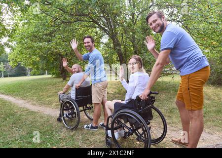 Amici allegri, due dei quali stanno usando le sedie a rotelle, sventolando e sorridendo in un parco verde lussureggiante. Raffigurante l'inclusione e l'amicizia in un ambiente naturale. Foto Stock