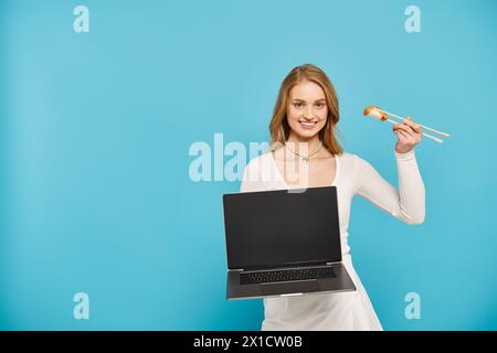Una donna bionda che tiene in mano un laptop e cibo asiatico, che mette in mostra una miscela di tecnologia e delizie culinarie. Foto Stock