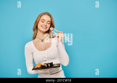 Un'elegante donna bionda regala un vassoio di sushi e bacchette, pronta a viziarsi con una deliziosa esperienza culinaria asiatica. Foto Stock