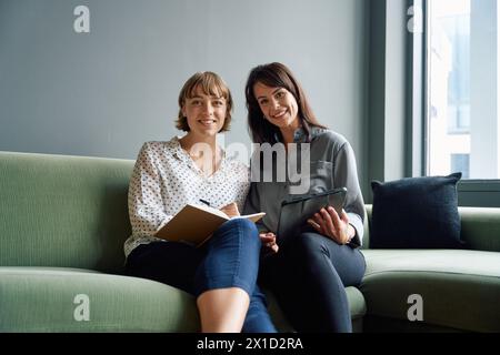 Due donne d'affari felici sedute sul divano utilizzando un tablet digitale durante una riunione in ufficio Foto Stock