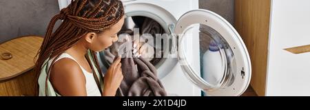 Una donna afroamericana con trecce afro guarda il suo riflesso in uno specchio mentre fa il bucato in un bagno. Foto Stock