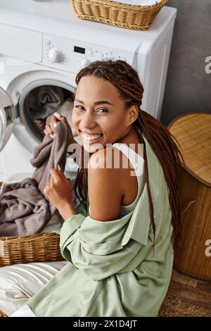 Una donna afroamericana con trecce afro siede sul pavimento accanto a una lavatrice, facendo il bucato in bagno. Foto Stock
