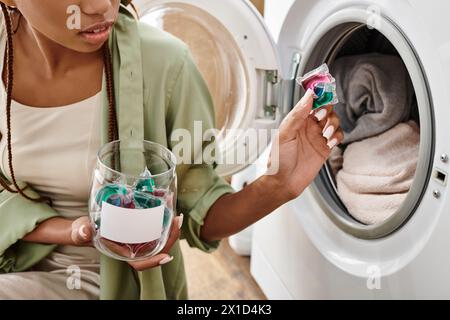 Una donna afro-americana con trecce afro gode di un momento tranquillo, tenendo una capsule di gel accanto a una lavatrice in un bagno accogliente. Foto Stock