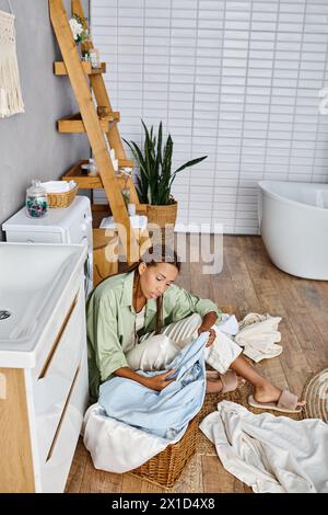 Una donna afroamericana con trecce afro siede sul pavimento del bagno, circondata da lavanderia mentre fa i lavori domestici. Foto Stock
