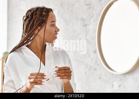 Donna afroamericana con dreadlock in accappatoio, che si ammira nello specchio del suo bagno moderno. Foto Stock