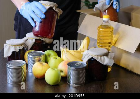 Un volontario che indossa guanti protettivi imballa il cibo in una scatola. Servizi di fornitura di generi alimentari durante la pandemia di coronavirus Foto Stock