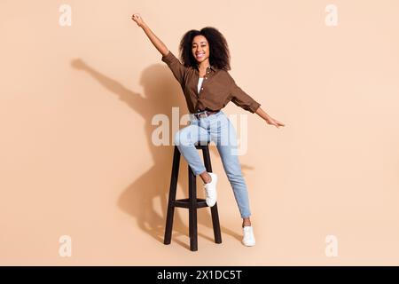 Foto a grandezza naturale di una camicia marrone vestita da donna di buon umore seduta su una sedia alzando il pugno in uno spazio vuoto isolato su sfondo di colore beige Foto Stock