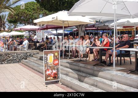 Los Cristianos, Tenerife, 16 aprile 2024 - i turisti bevono in bar sulla spiaggia in stile inglese, dove una pinta di birra costa 1,50 euro. - I turisti passano davanti ai manifesti Anti-Tourist lungo la spiaggia di Los Cristianos. Il poster recita che "le Isole Canarie hanno un limite” i cartelli gialli pubblicizzano la protesta anti-turistica di sabato 20 aprile. A Santa Cruz de Tenerife. Anche il parcheggio è un grosso problema nella zona, poiché orde di visitatori riempiono migliaia di spazi, causando mal di testa sia per i residenti che per i visitatori. Credito: Interrompi stampa Media/Alamy Live News Foto Stock