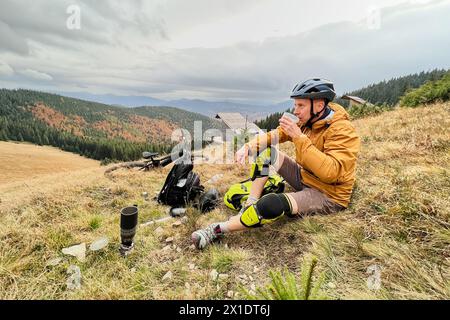 Uomo ciclista che beve una tazza con tè, indossa il casco da bici, siede sull'erba in montagna, riposa dopo aver guidato la bici elettrica. Mountain e-bike stesa a terra accanto a lui. Foto Stock