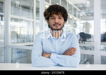Ritratto di un giovane uomo sicuro di sé con i capelli ricci, vestito con una camicia azzurra, seduto in un ambiente d'ufficio ben illuminato. Le braccia incrociate, trasuda professionalità e disponibilità. Foto Stock