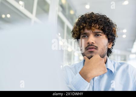 Un giovane uomo dai capelli ricci vestito con una camicia blu, che riflette profondamente mentre è seduto in un ambiente d'ufficio luminoso e contemporaneo. La sua espressione riflette la concentrazione e la contemplazione. Foto Stock