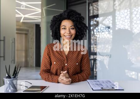 Giovane donna professionista con capelli ricci, con una camicia punteggiata, si impegna direttamente con la fotocamera in un ambiente luminoso e moderno. Foto Stock