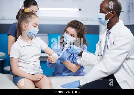Un medico afroamericano controlla e tratta la ferita di una ragazza caucasica in una clinica. Un medico di sesso maschile con un camice da laboratorio è assistito da un'infermiera in scrub blu con l'applicazione di una garza sul paziente. Foto Stock