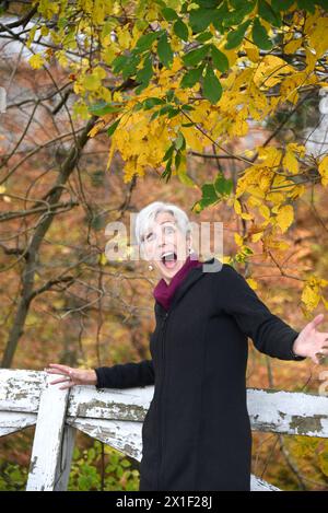 La donna anziana esclamò con gioia il colore autunnale oltre a una recinzione rustica in legno. Ha un cappotto nero e un maglione viola. Foto Stock