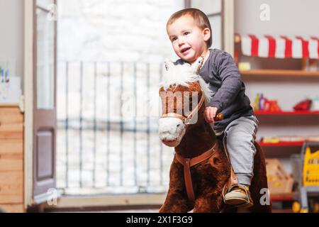 Bambino che cavalca sul suo cavallo a dondolo mentre gioca nella sala giochi di casa. Sullo sfondo si vedono le porte del balcone aperte. Foto Stock