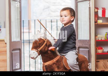 Bambino di due anni che cavalca sul suo cavallo a dondolo e tiene una spada di legno in una mano. Foto Stock