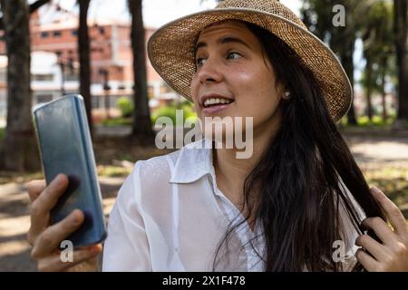 In primavera la giovane donna latinoamericana (33) è in un parco e usa il suo cellulare per effettuare una videochiamata. Concetto e tecnologia primaverili. Foto Stock