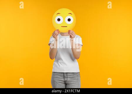 Donna che copre il viso con emoticon sorpresa su sfondo giallo Foto Stock