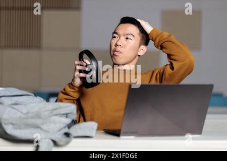 Un giovane studente universitario asiatico sta studiando e facendo i compiti alla sua scrivania, ma si sta incastrando, confuso e perso Foto Stock