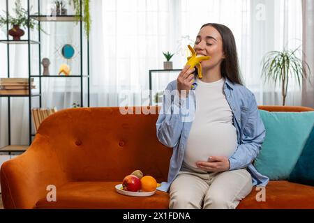 Una donna felice incinta con grande appetito sbuccia e mangia frutta di banana siede sul divano nell'interno del soggiorno. Assistenza all'infanzia per la maternità. Sorridente ragazza caucasica signora sceglie giusto cibo sano Foto Stock