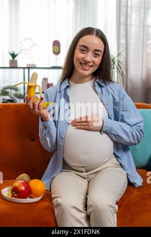 Una donna incinta felice con grande appetito sbuccia e mangia frutta di banana siede sul divano del soggiorno con interni moderni. Assistenza all'infanzia per la maternità. La ragazza sorridente sceglie il giusto cibo sano. Verticale Foto Stock