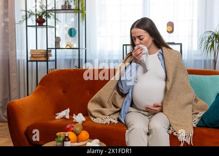 Donna caucasica incinta malsana ragazza avvolta in coperta che soffia il naso in tessuto mentre si siede sul divano divano in soggiorno. Mamma futura malata sensazione stanca di congestione nasale e sintomi di freddo a casa Foto Stock