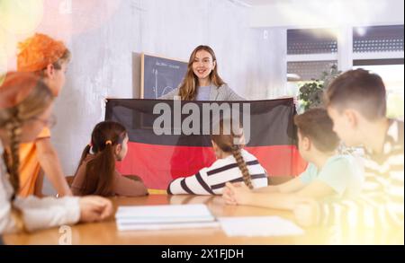 Gli scolari ascoltano attentamente la storia del paese Germania. L'insegnante tiene in mano la bandiera della Germania Foto Stock