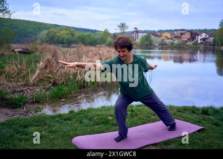 una donna anziana pratica yoga la mattina sulla riva del fiume, è venuta da casa in bicicletta Foto Stock