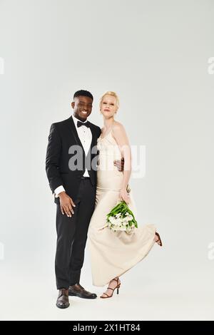 Una splendida sposa bionda in un abito da sposa e uno sposo afroamericano, entrambi in vestibilità formale, che posa elegantemente per un ritratto in uno studio su un grigio Foto Stock