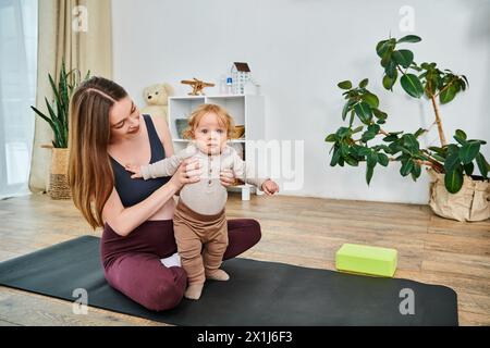 Una giovane madre si siede su una stuoia yoga che culla il suo bambino, guidata dal suo allenatore in un momento di pace connessione e cura. Foto Stock