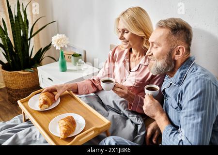 Una coppia matura in intimi casalinghi seduti su un divano, sorseggiando un caffè e viziandosi insieme con dolci in un'atmosfera calda e invitante. Foto Stock