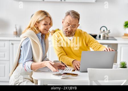 Una coppia matura e amorevole di articoli per la casa si siede a un tavolo, avvolto da uno schermo per laptop, e condivide un momento accogliente nella loro cucina di casa. Foto Stock